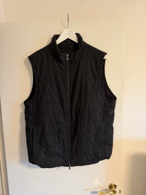 Oscar Jacobson Väst - Stilren mörkgrå dunväst med hög krage och hel dragkedja framtill. Västen har två sidofickor och quiltad design för extra värme. Passar perfekt över en hoodie eller tröja för en casual look.