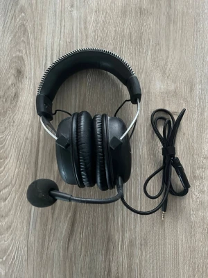 Svarta gaminghörlurar med mikrofon - Säljer ett par svarta over-ear gaminghörlurar med avtagbar mikrofon och kabel med 3,5 mm kontakt. Hörlurarna har vadderad bygel och mjuka öronkuddar i konstläder. Metallbågar på sidorna ger en cool look. Perfekta för gaming eller streaming.Är bara lite sönder i den muffen som sitter över mikrofonen men allt funkar perfekt!