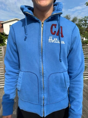 Blå zip hoodie från Hollister - Blå hoodie från Hollister med dragkedja, broderad röd CA och vit Hollister-text på bröstet. Mjuk insida, två stora fickor fram och justerbar huva med snören. Perfekt för chill dagar och streetwear-stil.