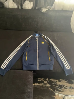 Blå Adidas zip hoodie med vita ränder - Mörkblå hoodie från Adidas med dragkedja, vita klassiska ränder längs ärmarna och gula detaljer vid fickor och axlar. Adidas-logga i gult på bröstet och ribbade muddar. Tillverkad i mjukt, stretchigt material som ger en sportig vibe.