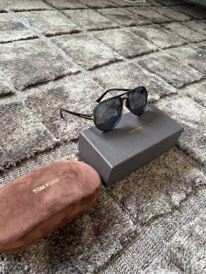 Svarta aviator-solglasögon Tom Ford - Snygga svarta aviator-solglasögon från Tom Ford med tunna metallbågar och mörka glas. Kommer med original brun sammetsfodral och ask med guldfärgad logga. Perfekta för dig som vill ha en klassisk och stilren look.