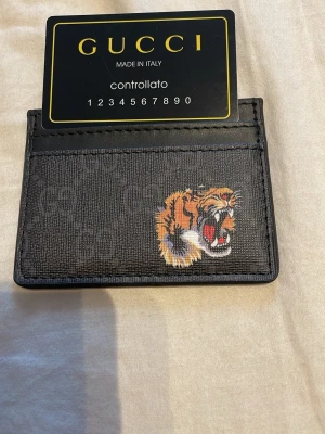 svart korthållare med tiger - Snygg svart korthållare i canvas och läder med diskret GG-mönster och en cool tigerbrodyr på framsidan. Perfekt för kort och ID, smidig att ha i fickan. Klassisk och trendig accessoar för dig som gillar lyxiga detaljer.