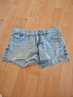 Ljusblå jeansshorts - Säljer ett par ljusblå jeansshorts med råa kanter och snygga broderade detaljer på bakfickorna. Shortsen har bälteshällor och två fickor bak med knappstängning. Perfekta för varma dagar!