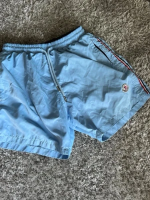 Ljusblå badbyxor från Moncler - Snygga ljusblå badbyxor från Moncler med elastisk midja och dragsko. Klassisk Moncler-logga på benet och rödblå rand längs sidan. Tillverkade i snabbtorkande material, perfekta för stranden eller poolen.