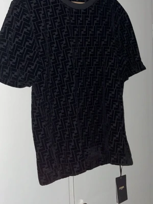 Svart mönstrad t-shirt från Fendi - Säljer en svart t-shirt från Fendi med det klassiska FF-mönstret över hela plagget. T-shirten har korta ärmar och rund halsringning. Materialet känns lyxigt och har en sammetsliknande finish som ger en exklusiv vibe.