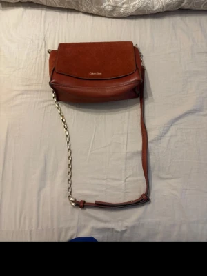 Röd axelväska från Calvin Klein - Snygg röd axelväska från Calvin Klein i en mix av slätt och mockaliknande material. Väskan har ett lock med logga, silverfärgad kedja på axelremmen och stilren design. Perfekt för dig som vill ha en trendig och praktisk väska.