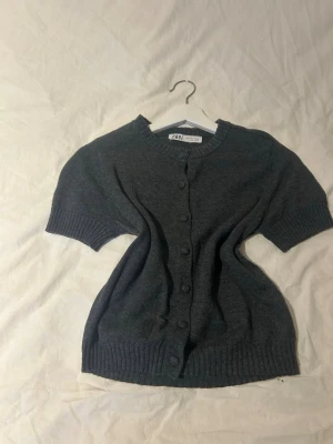 Mörkgrå kortärmad kofta från Zara - Kortärmad kofta från Zara i mörkgrått, med rund halsringning och knappar framtill. Ribbstickade detaljer vid ärmslut, hals och nederkant. Perfekt att slänga över en topp för en chill look.