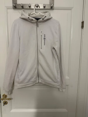 Vit hoodie från Sail Racing - Snygg vit hoodie från Sail Racing med dragkedja framtill och en bröstficka med zip. Märkets logga är tryckt på bröstet. Hoodien har huva med dragsko och två sidofickor. Perfekt för en avslappnad och sportig stil. Barn storlek 176, men är som S/M