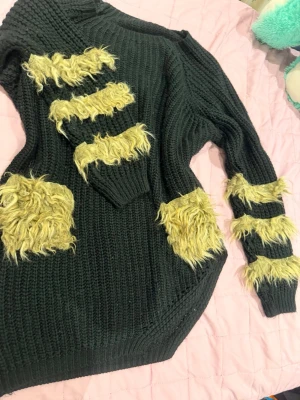 Svart stickad tröja med fluffiga detaljer - Unik svart stickad tröja med flera gula fluffiga partier på bröstet, ärmarna och ena höften. Oversized passform och grovstickad struktur ger en cool och trendig look. Perfekt för dig som vill sticka ut med din stil.