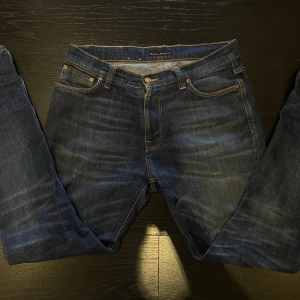 Mörkblå jeans från Nudie Jeans W30 L32 - Säljer ett par mörkblå jeans från Nudie Jeans med klassiska orange sömmar och snygga broderade bakfickor. Jeansen har rak passform och är tillverkade i slitstarkt denimtyg. 