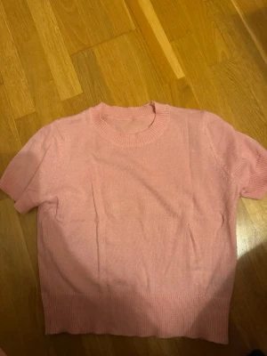 Ljusrosa stickad topp med kort ärm - Supersöt ljusrosa stickad topp med korta ärmar och rund halsringning. Toppen har ribbade detaljer vid hals, ärmslut och nederkant. Perfekt att styla med jeans eller kjol för en fräsch look.