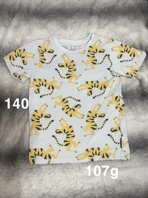Vit t-shirt med tigertryck - Säljer en vit t-shirt med coolt gult och svart tigertryck över hela plagget. T-shirten har rund hals och korta ärmar. Perfekt för dig som gillar djurmotiv och vill sticka ut lite extra.🌸 Fint använt skick. Storlek 140 