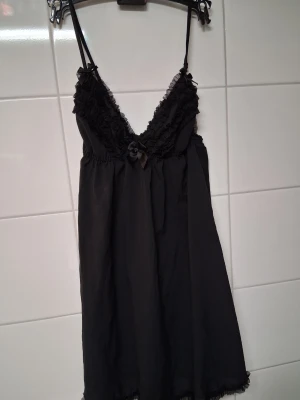 Svart nattlinne med spetsdetaljer - Snyggt svart nattlinne med tunna axelband och djup V-ringning. Dekorerat med volangkantad spets och en liten rosett framtill. Tillverkat i ett lätt och mjukt tyg som känns bekvämt mot huden. Perfekt för mysiga kvällar hemma.