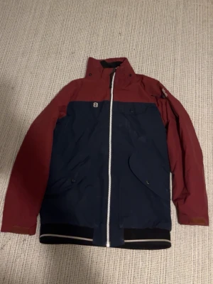 Blå och vinröd vindjacka med dragkedja - Snygg tvåfärgad vindjacka i marinblått och vinrött med hög krage och vit dragkedja framtill. Jackan har praktiska fickor med lock och muddar vid ärmslut och nederkant. Perfekt för dig som vill ha en stilren och funktionell jacka. Annonsen är ute på Vinted också ifall ni skulle se den där. 