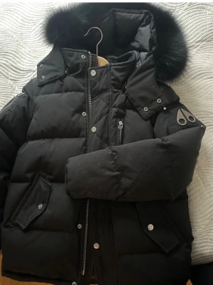 Svart dunjacka från Moose Knuckles - Snygg svart dunjacka från Moose Knuckles med pälskantad huva och flera fickor. Jackan har quiltad design, dragkedja och knappar framtill samt ikonisk logga på ärmen. Perfekt för kalla vinterdagar.