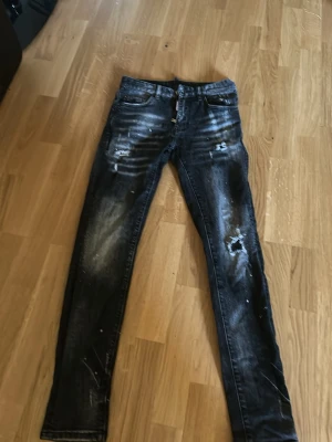 Svarta slitna jeans från Dsquared2 - Säljer ett par svarta  jeans från Dsquared2 med slitningar, hål och vitmålade detaljer. Jeansen har klassisk femficksmodell, knappgylf och en cool tvättad look. Perfekta för dig som gillar streetstyle och vill sticka ut.