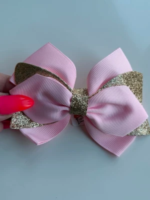Rosa hårrosett med glitterdetaljer - Söt hårrosett i ljusrosa tyg med glittriga gulddetaljer. Rosetten har dubbla lager och fästs enkelt med en metallklämma på baksidan. Perfekt accessoar för att piffa upp frisyren med lite extra bling.