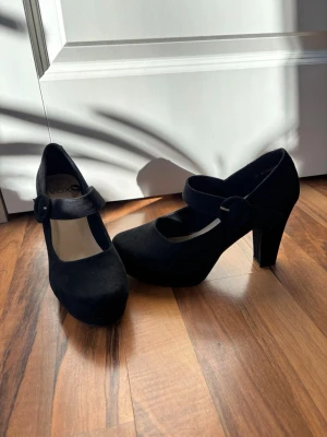 Svarta Mary Jane-pumps från Vox - Svarta pumps från Vox med rund tå och bred klack. Skorna har en bred rem över foten och är tillverkade i mockaimitation. Väldigt bekväma! Tecken på användning kan förekomma !