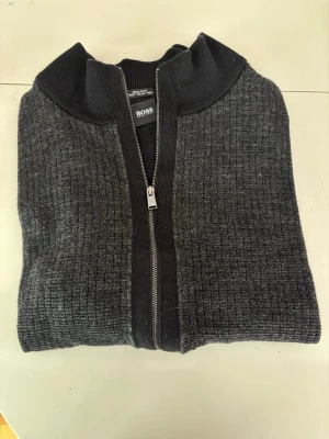 Mörkgrå ziptröja från BOSS - Snygg mörkgrå ziptröja från Hugo BOSS med ribbad krage och strukturstickat mönster. Tröjan har hel dragkedja framtill och breda muddar.