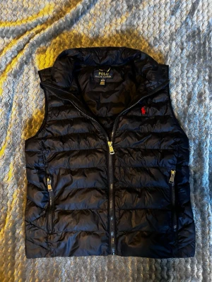 Svart dunväst från Polo Ralph Lauren - Snygg svart dunväst från Polo Ralph Lauren med klassisk röd logga på bröstet. Västen har två dragkedjeförsedda fickor och hög krage. Tillverkad i glansigt material med quiltad design och gulddetaljer på dragkedjorna. Perfekt för lager på lager-stil.