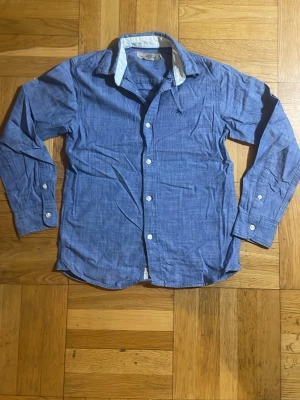 Blå långärmad skjorta med vita detaljer - Snygg blå skjorta med vita knappar och kontrasterande vit insida på kragen. Skjortan har klassisk krage och lång ärm, perfekt för en clean och avslappnad look. Tillverkad i mjukt bomullstyg som känns skönt mot huden.