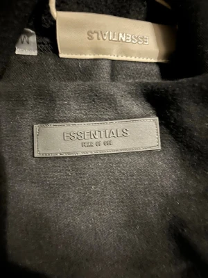 Svart hoodie från Essentials Fear of God - Säljer en svart hoodie från Essentials Fear of God med diskret logga på bröstet, etikett i nacken och ennu en logga på nacksidan. Hoodien har en klassisk passform och är tillverkad i mjukt material som känns riktigt skönt mot huden. Perfekt för dig som gillar streetwear och stilrena plagg.