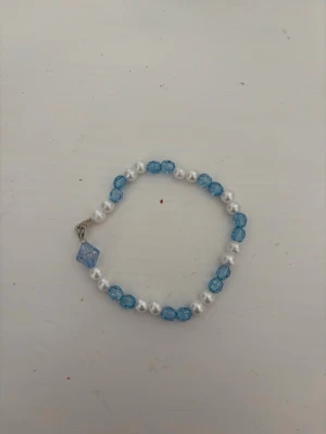 Blått och vitt pärlarmband - Snyggt armband med växelvis blå och vita pärlor. Armbandet har en liten blå kristallformad pärla som hänge. Perfekt accessoar för att addera lite färg till din outfit.