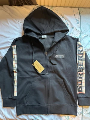 Svart Burberry hoodie med dragkedja - Snygg svart hoodie från Burberry med dragkedja och klassiska rutiga detaljer längs ärmarna. Stor Burberry-logga på ena ärmen och mindre logga på bröstet. Utrustad med huva och känguruficka framtill. Perfekt för en trendig och avslappnad stil.