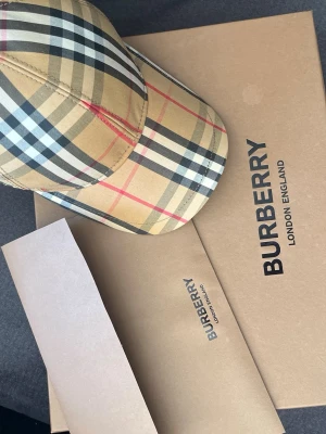 Burberry klassisk rutmönstrad keps - Säljer en snygg keps från Burberry med ikoniskt beige rutmönster och inslag av svart, vitt och rött. Kepsen har böjd skärm och justerbar rem baktill. Perfekt för dig som vill ha en trendig accessoar med lyxig känsla. Finns äkthetsbevis kan tänka mig sänka pris 