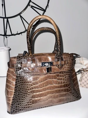 Brun krokopräglad handväska - Säljer en brun handväska i krokopräglat konstläder med silverfärgade metalldetaljer och lås framtill. Väskan har två handtag och en avtagbar axelrem. Klassisk och strukturerad modell med lyxig känsla och snygg finish.