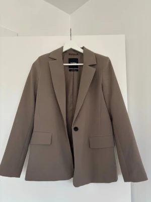 Beige kavaj från Pull&Bear - Stilren beige kavaj från Pull&Bear med klassisk passform och en knapp framtill. Kavajen har två fickor och är tillverkad i ett mjukt material. Perfekt för en stilren look! Klänningen har använts - men har inga synliga slitage förutom trådar från öppningen i ryggen som man kan öppna.