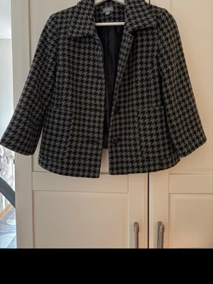 Svartvit kort kappa med hundtandsmönster - Stilren svartvit kappa med klassiskt hundtandsmönster och bred krage. Modellen är kortare med trekvartsärmar och knäppning framtill. En defekt på ryggen där tyget ”glidit isär”