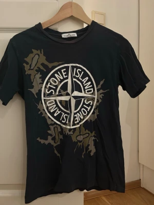 Stone Island Junior camo t-shirt - Snygg mörkblå t-shirt från Stone Island Junior med stort vitt logotryck och camo-detaljer i grönt och beige på framsidan. Klassisk rund hals och korta ärmar. Perfekt för dig som vill sticka ut med en streetig vibe. Lite blekt men annars bra kvalité 