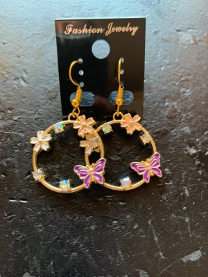 Guldiga örhängen med blommor & fjärilar - Snygga örhängen i guldfärgad metall med rund form, dekorerade med rosa blommor, lila fjärilar och glittrande stenar. Perfekta för att ge din look en söt och färgglad touch. Hängande modell med krok.