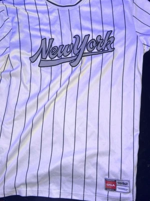 Vit randig New York t-shirt - Snygg vit t-shirt med svarta ränder och texten 'New York' i grått framtill. Klassisk basebollstil med rund hals och korta ärmar. Märkt med USA 1992 nere vid fållen. Perfekt för en sportig och avslappnad look.