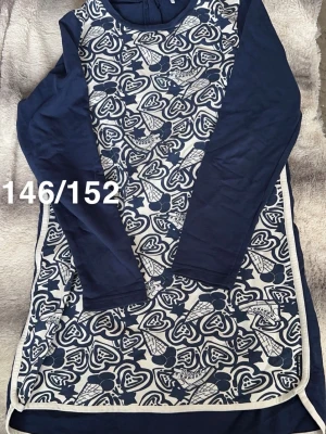 Marinblå och vit långärmad klänning - Klänning i marinblått och vitt med lång ärm och hjärtmönster framtill. Klänningen har kontrasterande vita kanter. Perfekt för dig som gillar lekfulla detaljer och bekväm passform.🌸 Sparsamt använd. Storlek 146/152. 