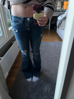 Blå bootcut jeans med slitningar - Säljer ett par blå bootcut jeans med slitningar på knäna och snygga broderade detaljer på bakfickorna. Jeansen har låg midja, vita kontrastsömmar och klassisk femficksmodell. Perfekta för en avslappnad och trendig look. Är i xs men passar för S också 