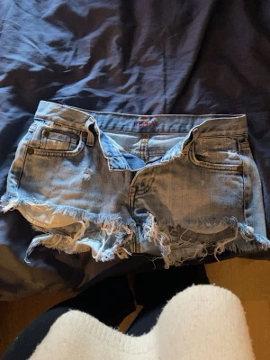 Blå low waisted jeansshorts med fransar från Replay - Säljer ett par ljusblå jeansshorts från Replay med råa, fransiga kanter och slitna detaljer. Klassisk femficksmodell med dragkedja och knapp. Perfekta för sommaren och ger en avslappnad vibe. Har ett hål som är lagat längst ner på rumpan men inget som syns på långt håll 💞