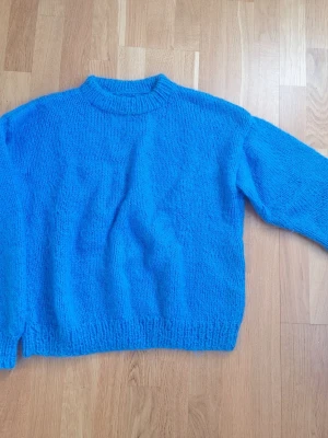 Blå hemstickad tröja i lurvigt garn - En klarblå hemstickad tröja med rund hals och långa ärmar. Tröjan har ribbstickade muddar vid ärmslut och nederkant. Stickat i gosigt sysntetfarn som går bra att tvätta i maskin och ger en mysig känsla.