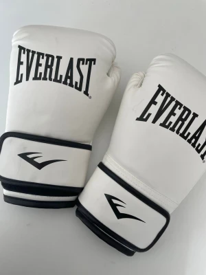 Vita boxningshandskar Everlast - Säljer ett par vita boxningshandskar från Everlast med svart logga och detaljer. Handskarna har bred kardborrestängning runt handleden för extra stöd och är tillverkade i syntetmaterial. Perfekta för träning och kampsport.