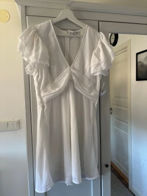 Vit kortklänning med volangärmar - Söt vit kortklänning med v-ringning och dubbla volangärmar. Klänningen har en omlottliknande detalj framtill. Lapp kvar.