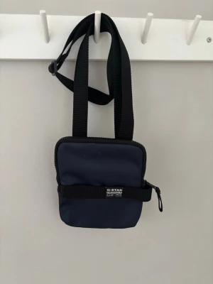 Mörkblå axelväska från G-Star RAW - Snygg mörkblå axelväska från G-Star RAW med justerbar svart axelrem. Väskan har en fyrkantig form, dragkedja upptill och en svart rem med logotyp på framsidan. Tillverkad i slitstarkt tyg, perfekt för att bära det viktigaste.
