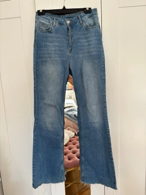 Blåa utsvängda jeans med rå kant - Säljer ett par ljusblå jeans med hög midja och utsvängda ben. Jeansen har råa, fransiga benslut och klassisk femficksdesign. Materialet är denim i bomull och de har en snygg tvättad look. Perfekta för en trendig och avslappnad stil.