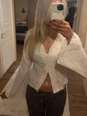 Vit spetsblus med vida ärmar - Säljer en superfin vit spetsblus med v-ringning och pärlknappar framtill. Blusen har långa, vida ärmar och ett lätt figurnära snitt. Materialet är spets med blommigt mönster som ger en romantisk vibe. Perfekt att styla med jeans eller kjol. Toppen är aldrig använd.