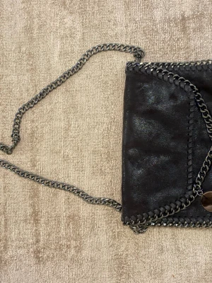 Svart Stella McCartney  liknade axelväska - Snygg svart axelväska som liknar  Stella McCartney med kedjerem och dekorativa kedjedetaljer runt kanterna. Väskan har en kuvertformad design och ett metallmärke framtill. Tillverkad i veganskt läder som ger en cool glansig finish. Har aldrig använt väskan !💖💖 köpte den på en marknad i Spanien så det är inget märke på den!💞💞