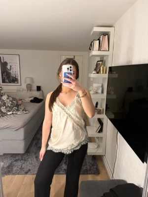 Krämfärgat linne med spetsdetaljer - Säljer ett krämfärgat linne med tunna axelband och fina spetsdetaljer i vitt längs ringningen och nederkanten. Aldrig använt