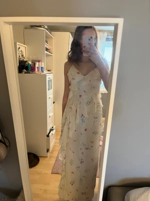 Vit långklänning med blommigt mönster - Säljer en vit långklänning med tunna axelband och v-ringning. Klänningen har ett blommigt mönster i pastellfärger och volangdetaljer framtill. Perfekt för sommarens alla tillfällen! Materialet känns lätt och luftigt.