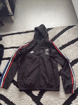 Svart Real Madrid adidas jacka - Svart sportjacka från adidas med Real Madrid-logga på bröstet. Jackan har huva, dragkedja och fickor samt ränder i rött, vitt och blått längs ärmarna. Tillverkad i lätt syntetmaterial, perfekt för träning eller vardag.