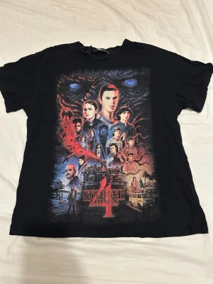 Stranger Things svart t-shirt - Cool svart t-shirt med stort Stranger Things-tryck på framsidan. Motivet visar flera karaktärer och detaljer från serien. T-shirten har rund hals och korta ärmar. Perfekt för dig som älskar retro och popkultur. Storlek S men passar även mig som är M.