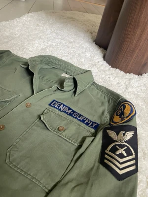 Grön skjorta från Ralph Laurent  - Säljer en militärinspirerad grön skjorta från Ralph Laurent  med coola patchar på ärmen och bröstet. Skjortan har två bröstfickor med knappar och klassisk krage. Perfekt för dig som gillar utility-stil och detaljer.